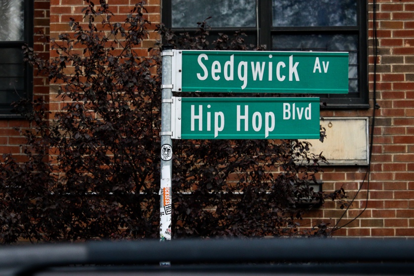 A signpost with "Sedgwick Av" on top of "Hip Hop Blvd". Windows on a brick building and a tree with dark leaves are seen in the background.